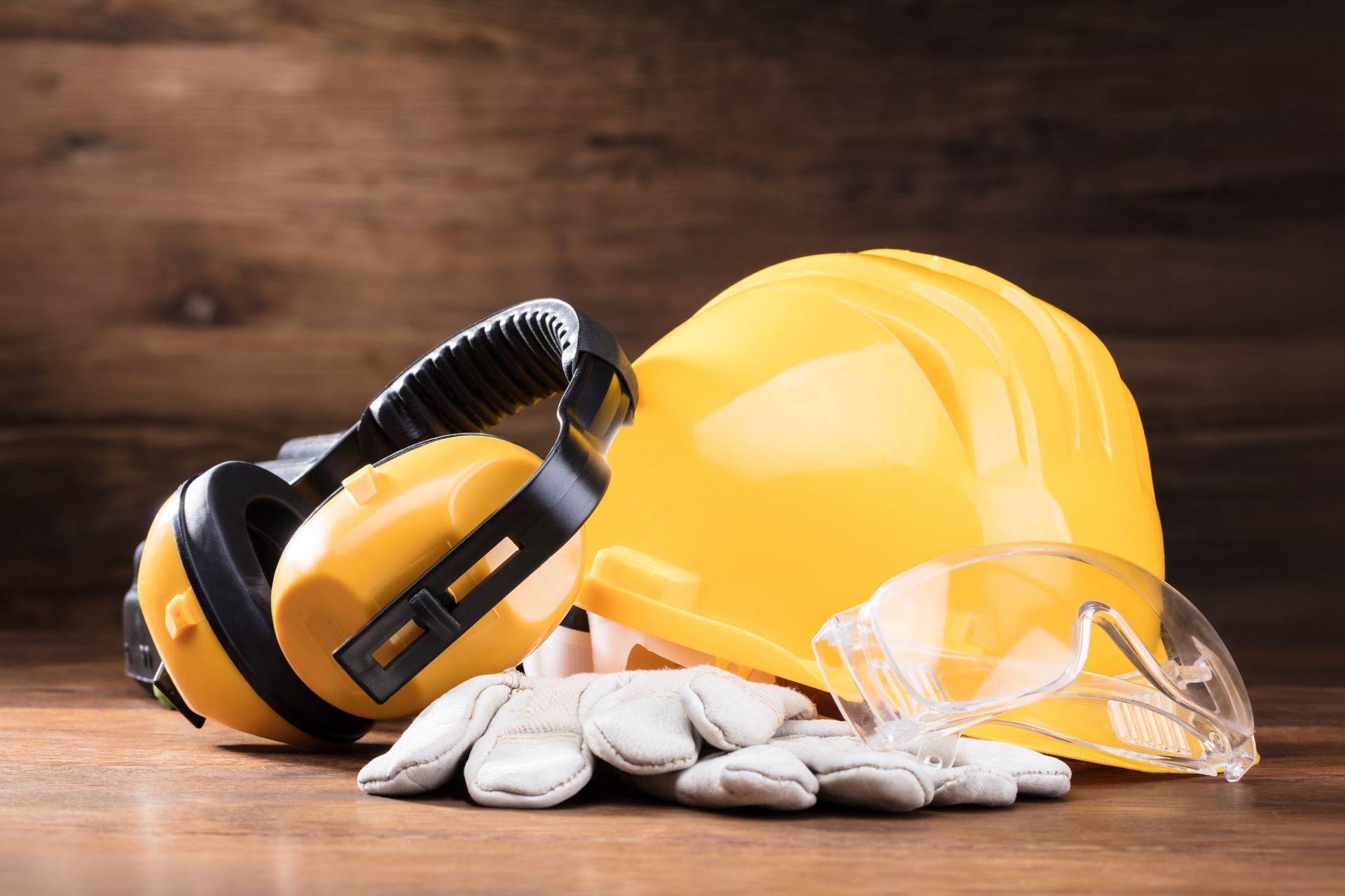 Yellow hard hat, ear muffs, safety glasses, and gloves on wooden surface. Safety equipment.