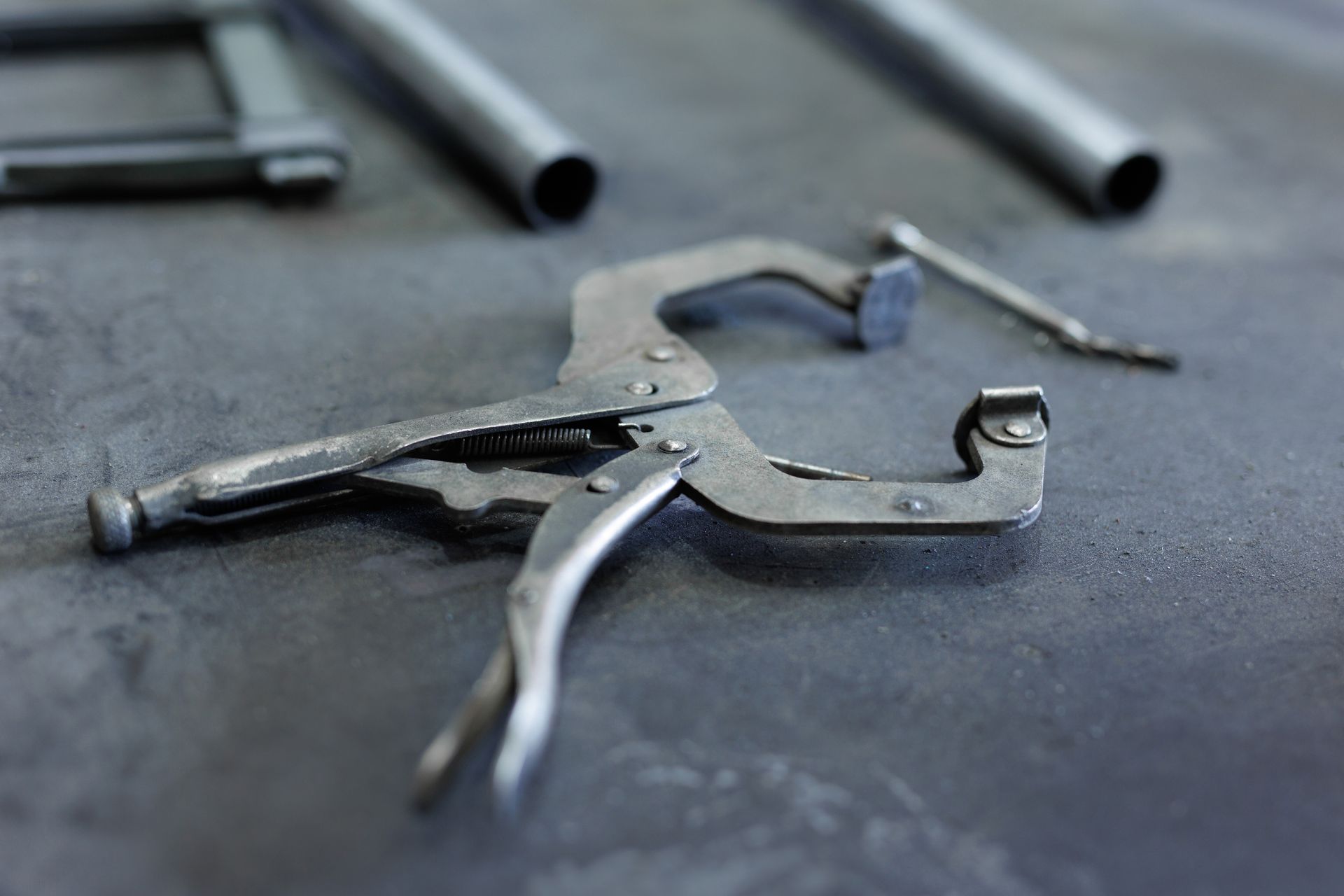 Metal locking pliers next to metal tubes on a dark surface.